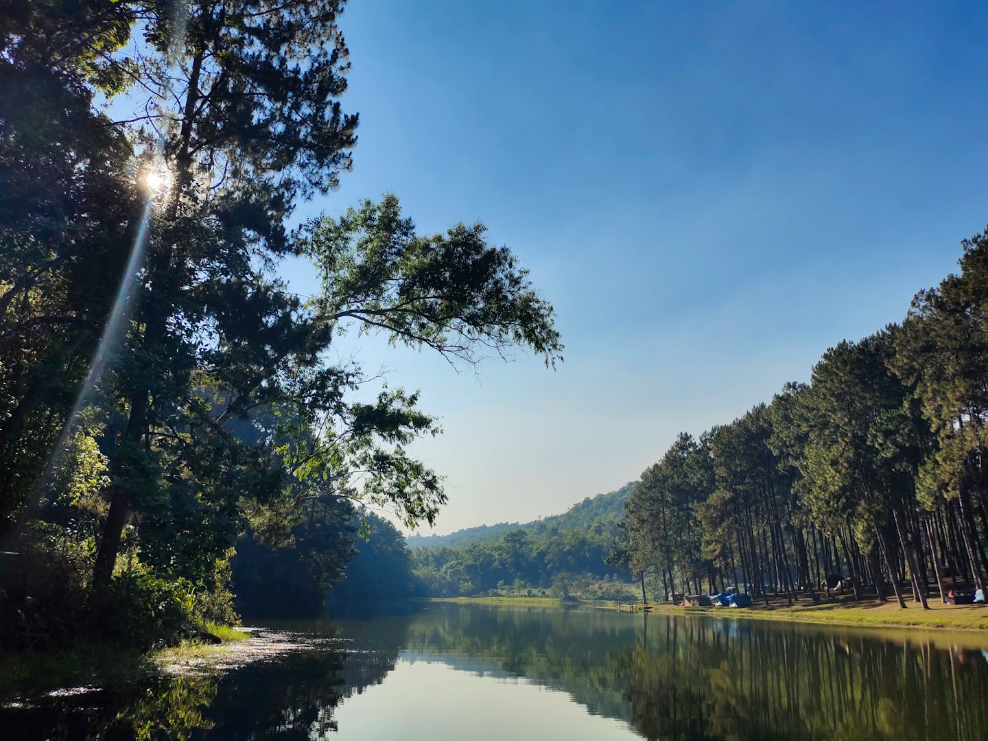 Calm water reflects the surrounding forest and distant green hills.